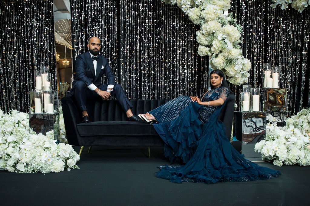Man in a tuxedo and a woman in a navy gown recline on a black sofa at a formal event, surrounded by white flowers and candles.