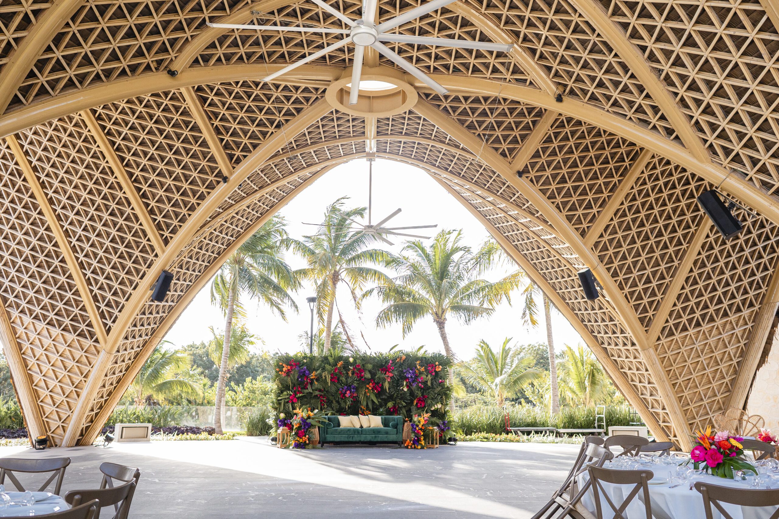 Open-air wooden lattice pavilion interior with large white ceiling fans, palm trees beyond, and colorful floral arrangements around a seating area for an event.