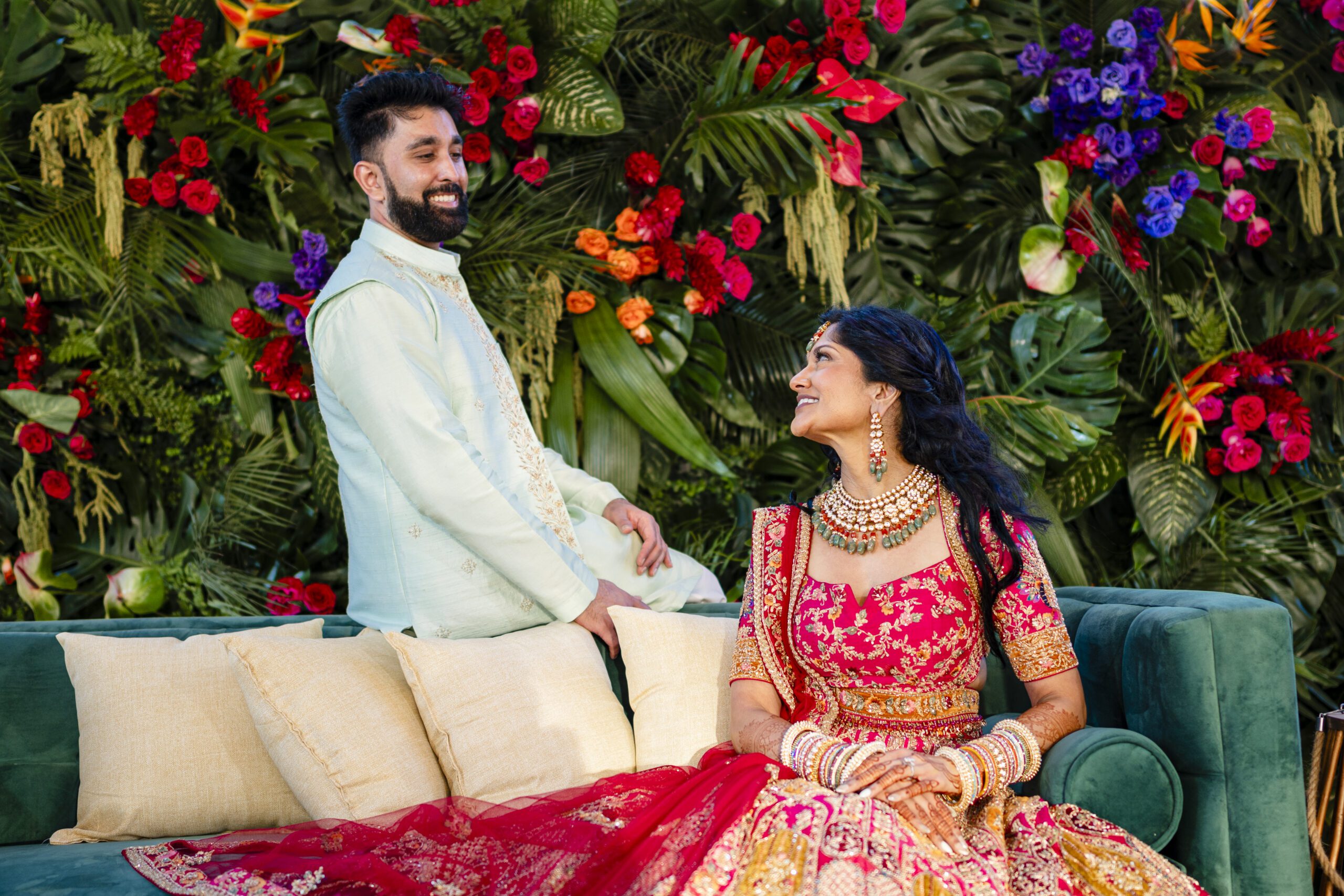 Indian bride and groom in traditional attire posing together in front of a vibrant flower wall.
