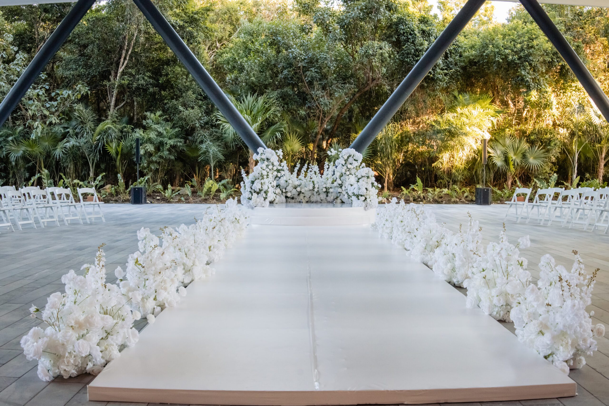 Wedding ceremony aisle with white floral arrangements lining a glossy white runway and rows of chairs, set against a lush green garden backdrop.