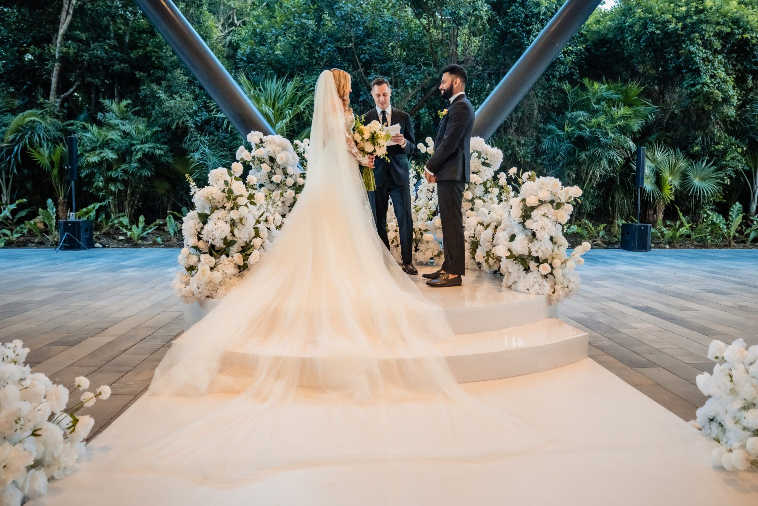 Bride in a long white gown and veil exchanges vows with the groom as the officiant reads, surrounded by white floral arrangements outdoors.