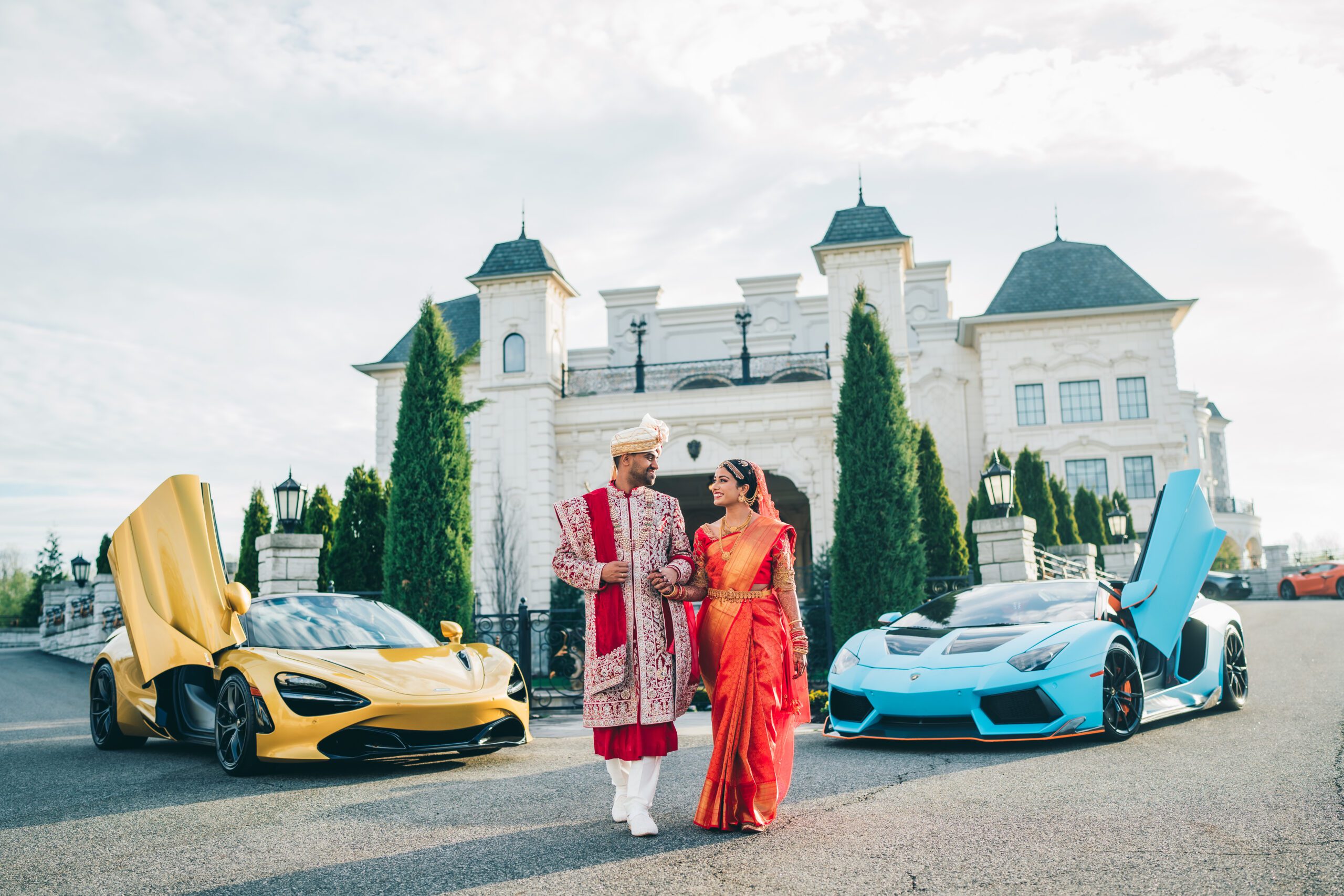 Indian bride and groom in traditional red and cream attire pose in front of a white mansion, with a yellow and a blue luxury sports car nearby.