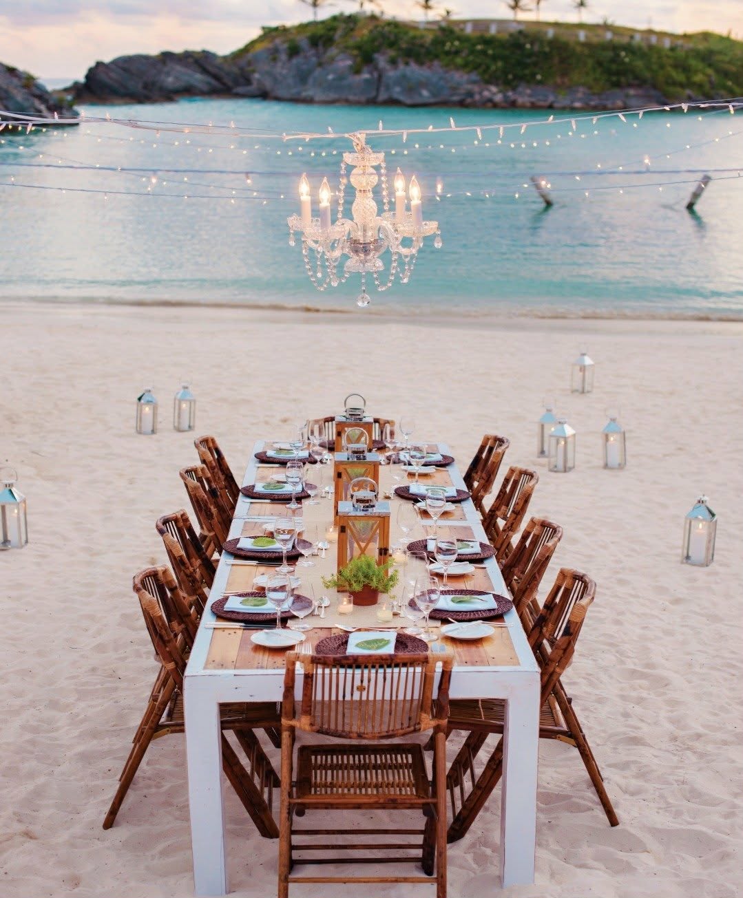Long wooden dining table set for a beachside dinner on the sand, with a hanging crystal chandelier and lanterns, facing turquoise sea.